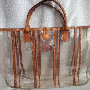May include: A clear plastic tote bag with brown leather trim and straps. The bag has a striped design and a small leather tag with a logo.