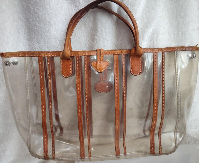 May include: A clear plastic tote bag with brown leather trim and straps. The bag has a striped design and a small leather tag with a logo.
