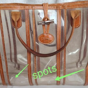 May include: Clear plastic tote bag with brown leather trim and straps. The bag has a striped design and a metal clasp closure. There are spots on the bag.