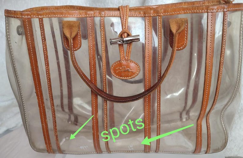 May include: Clear plastic tote bag with brown leather trim and straps. The bag has a striped design and a metal clasp closure. There are spots on the bag.