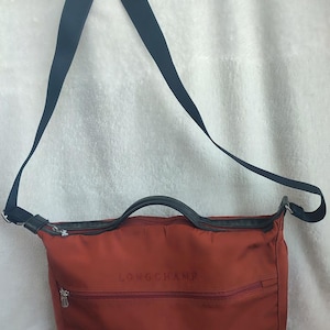 Op de afbeelding: Een rode Longchamp schoudertas met een zwarte band en een bovenste handvat. De tas heeft een ritsvak aan de voorkant en het Longchamp-logo is geborduurd op de voorkant.
