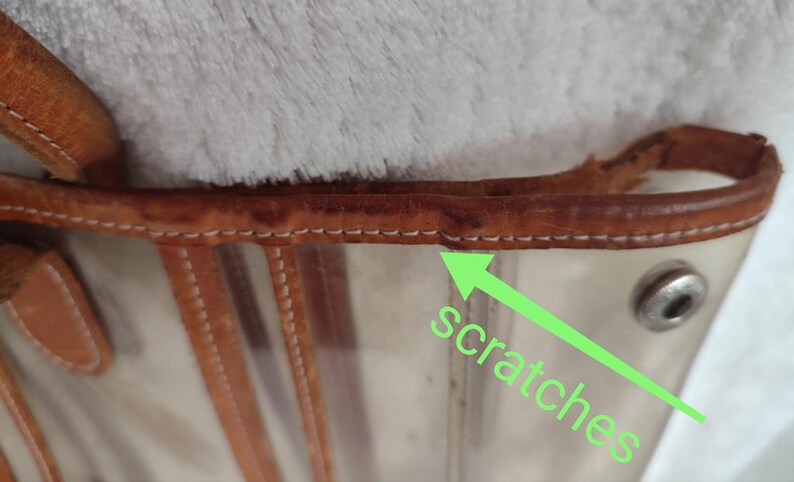 May include: Close-up of a clear plastic tote bag with brown leather trim. The leather trim has visible scratches.