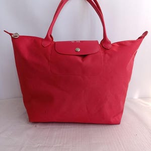 May include: A bright red tote bag with matching handles and a flap closure. The bag features a silver-toned zipper and a small flap with the initials "WM". The bag is made of a durable material and is designed for everyday use.