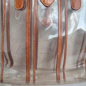 May include: A clear plastic tote bag with brown leather straps and stitching. The bag has a striped design.