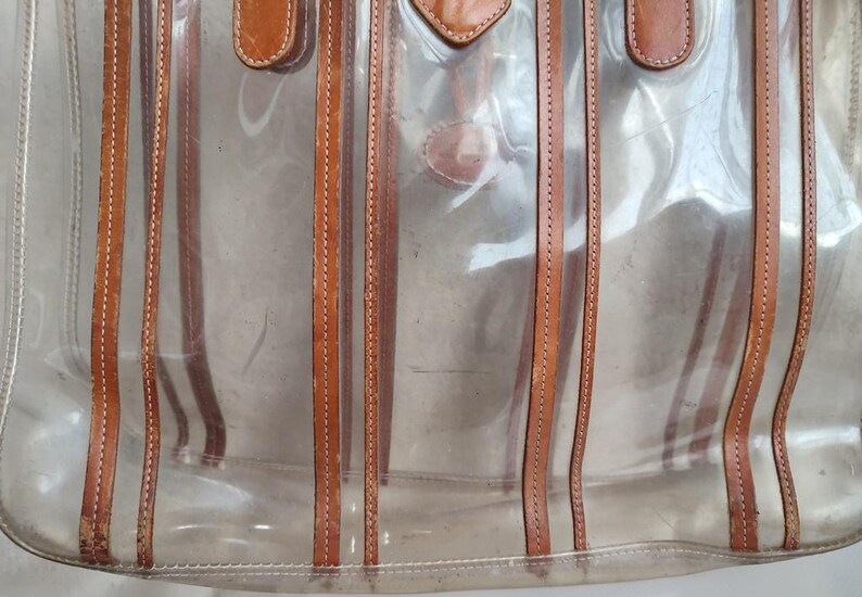 May include: A clear plastic tote bag with brown leather straps and stitching. The bag has a striped design.