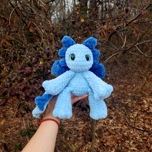 May include: A crocheted blue dragon plush toy with sparkly green eyes. The dragon is sitting up with its wings out and its tail curled around its legs.