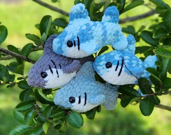 May include: Three crocheted shark plushies in blue and gray colors. The sharks are arranged in a cluster on a green leafy branch.