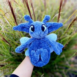 May include: A blue crocheted plush toy with large eyes and wings. The toy is made of a soft, fuzzy material and has a whimsical, friendly expression.