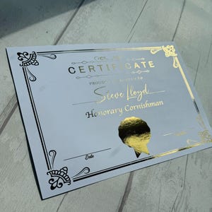May include: A white certificate with gold foil lettering and decorative borders. The text reads CERTIFICATE, PROUDLY PRESENTED TO, Steve Lloyd, Honorary Cornishman, and Date. A gold foil seal is positioned in the lower right section of the document.