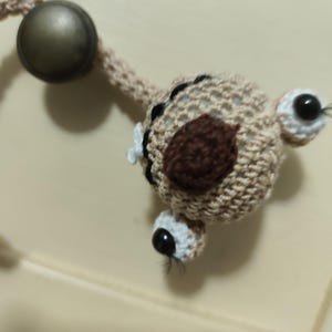 Puede incluir: Una criatura beige tejida con ojos negros y blancos, una mancha marrón y una pequeña flor blanca. La criatura está unida a un cordón beige tejido y a una bola de metal oscuro.