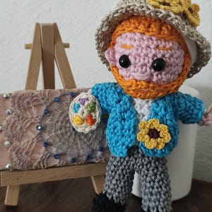 Könnte beinhalten: Handgefertigte Häkelpuppe, die Vincent van Gogh darstellt, mit einer blauen Jacke, grauen Hosen und einem Hut mit gelben Blumen. Die Puppe hält eine Miniaturpalette und steht neben einer kleinen Staffelei mit einem Gemälde.