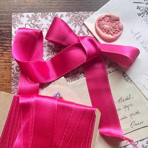 Op de afbeelding: Een helder fuchsia satijnen lint, in een strik geknoopt en opgerold, wordt getoond met vintage briefpapier. De scène omvat enveloppen en papier met bloemmotief. Twee waszegels voegen een vleugje elegantie toe aan de opstelling.