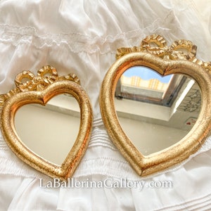 Peut inclure: Deux miroirs en forme de cœur dorés avec des détails de ruban doré ornés. Les miroirs sont sur un fond blanc.
