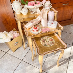 K&ouml;nnte beinhalten: Arrangement von Ballettthemen auf goldenen Beistelltischen. Auf den Tischen befinden sich rosa Spitzenschuhe, eine Teekanne, Tassen und eine dekorative Dose. Ein Korb mit rosa Blumen und ein kleiner Kuchen sind ebenfalls vorhanden. Die Website LaBallerinaGallery.com ist sichtbar.