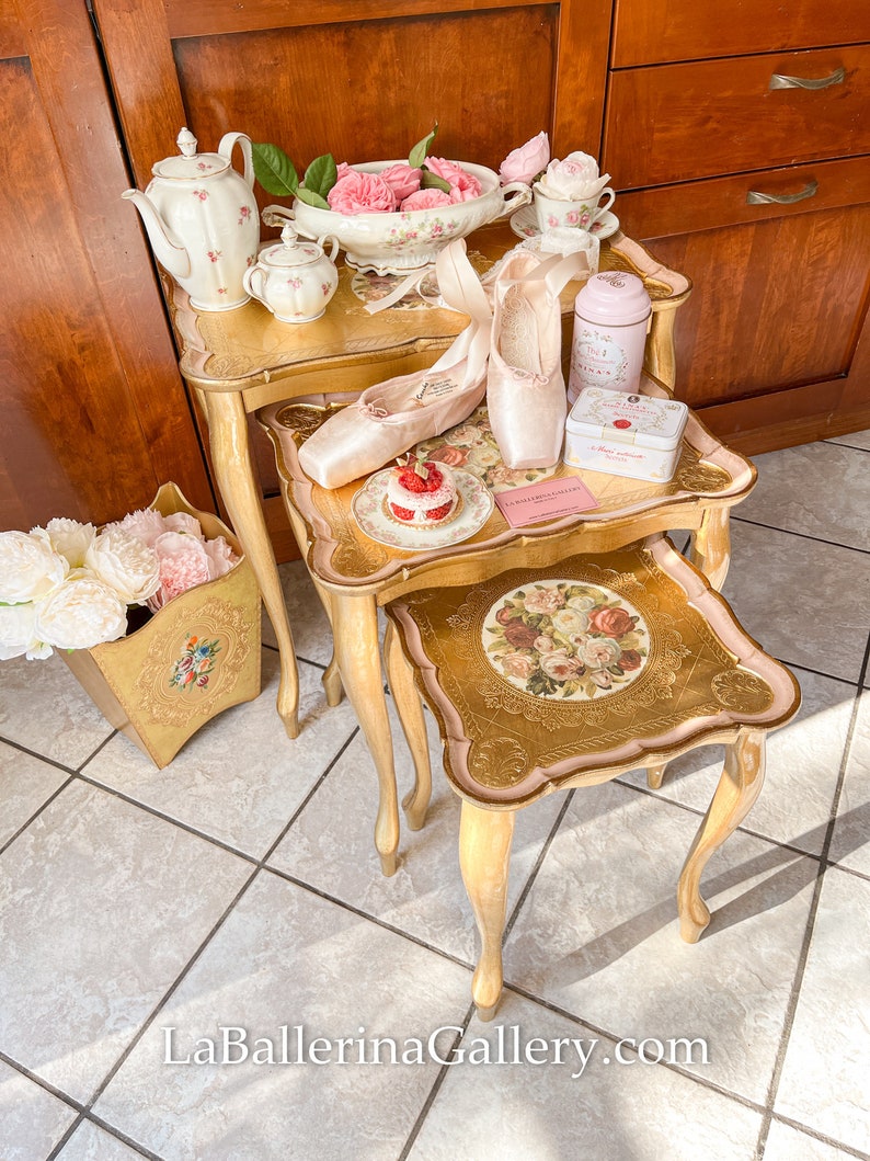 K&ouml;nnte beinhalten: Arrangement von Ballettthemen auf goldenen Beistelltischen. Auf den Tischen befinden sich rosa Spitzenschuhe, eine Teekanne, Tassen und eine dekorative Dose. Ein Korb mit rosa Blumen und ein kleiner Kuchen sind ebenfalls vorhanden. Die Website LaBallerinaGallery.com ist sichtbar.