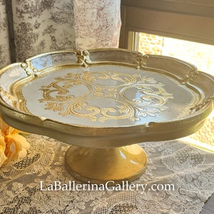 May include: A white cake stand with a gold rim and intricate gold design. The stand has a pedestal base.