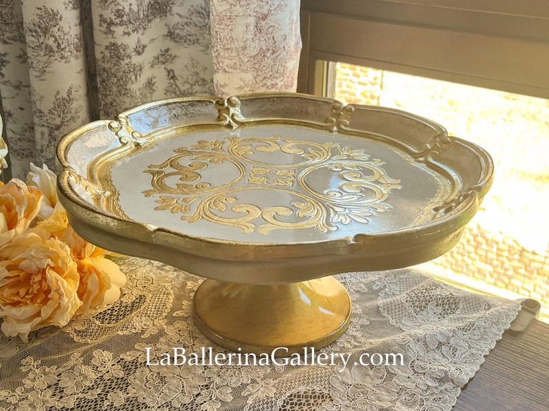 May include: A white cake stand with a gold rim and intricate gold design. The stand has a pedestal base.