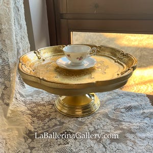 Puede incluir: Un soporte para tartas dorado con un diseño decorativo y ornamentado. El soporte tiene una parte superior redonda y una base de pedestal. Una pequeña taza de té blanca y un platillo están encima del soporte.