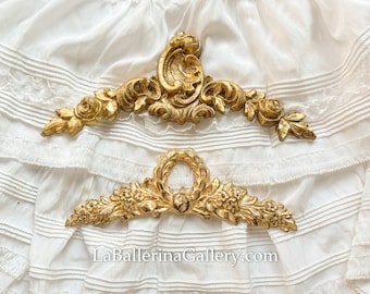Può includere: Due pezzi decorativi in metallo dorato con motivi floreali ornamentali. Il pezzo superiore è un design più grande e elaborato con un ricciolo centrale. Il pezzo inferiore è un design più piccolo e più simmetrico con un cerchio centrale.