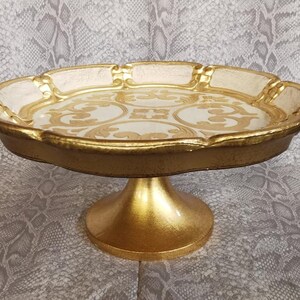 May include: A gold and white cake stand with a decorative design. The stand has a round top and a pedestal base.