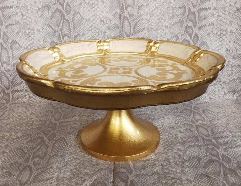 May include: A gold and white cake stand with a decorative design. The stand has a round top and a pedestal base.