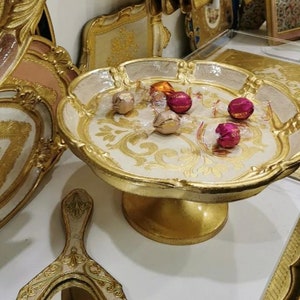 Puede incluir: Una bandeja decorativa dorada y blanca con un borde elevado y un diseño ornamentado. La bandeja está llena de chocolates envueltos en rosa y dorado.