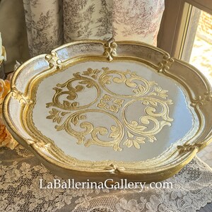 May include: A white and gold decorative tray with a raised, ornate, gold design. The tray has a scalloped edge and is made of wood.