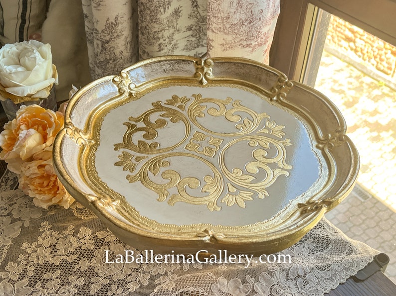 May include: A white and gold decorative tray with a raised, ornate, gold design. The tray has a scalloped edge and is made of wood.