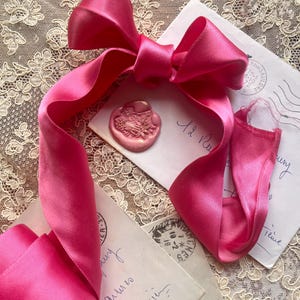 Può includere: Un nastro di raso rosa acceso, annodato in un grande fiocco, è adagiato su buste vintage e un sigillo di cera. Le estremità del nastro si estendono sulla scena. Le buste presentano scritte a mano e timbri postali.