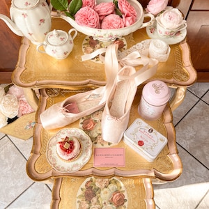 K&ouml;nnte beinhalten: Ein Stillleben mit Ballettschuhen, einer Teekanne, einer Teetasse und einem kleinen Kuchen auf einem dekorativen Tisch. Der Tisch ist mit floralen Mustern und Rosen verziert. Eine rosa Dose und eine Karte mit dem Text "La Ballerina Gallery" sind ebenfalls vorhanden.