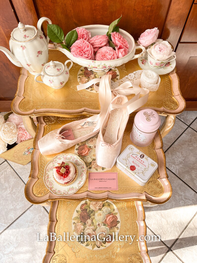 K&ouml;nnte beinhalten: Ein Stillleben mit Ballettschuhen, einer Teekanne, einer Teetasse und einem kleinen Kuchen auf einem dekorativen Tisch. Der Tisch ist mit floralen Mustern und Rosen verziert. Eine rosa Dose und eine Karte mit dem Text "La Ballerina Gallery" sind ebenfalls vorhanden.