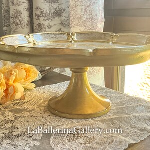 May include: A gold ceramic cake stand with a scalloped edge and a decorative design. The stand is on a white lace tablecloth.