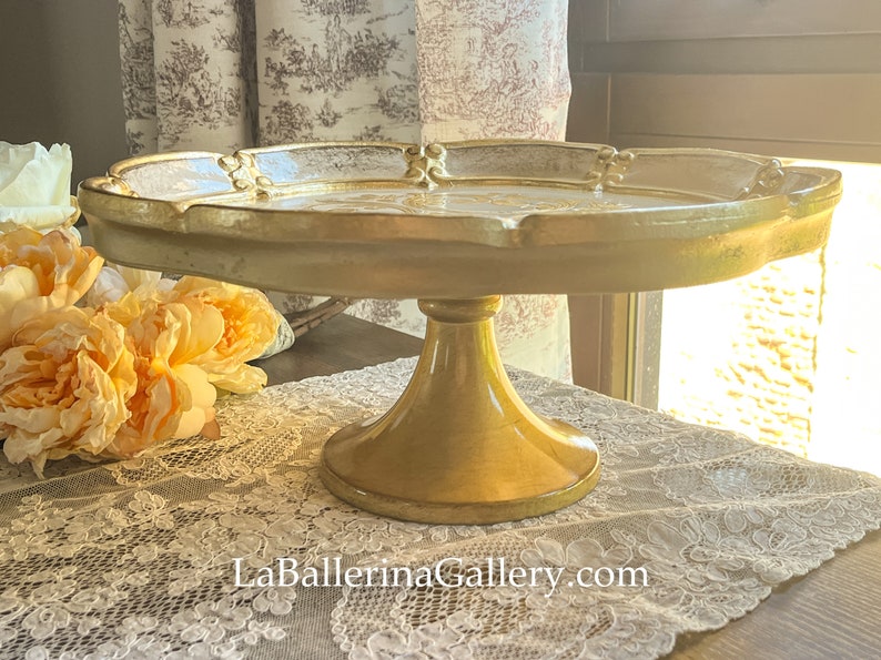 May include: A gold ceramic cake stand with a scalloped edge and a decorative design. The stand is on a white lace tablecloth.