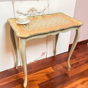 May include: A small, antique-style side table with a patterned top and curved legs. The table has a light green painted finish with a decorative border. A teacup and saucer sit on the table's surface. The website LaBallerinaGallery.com is visible.
