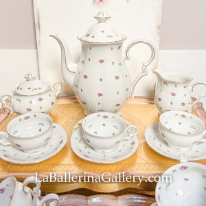 May include: A white porcelain tea set with a floral pattern of small pink roses and green leaves. The set includes a teapot, sugar bowl, creamer, and several teacups and saucers. Gold trim accents the edges.