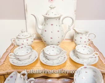 May include: A white porcelain tea set with a floral pattern of small pink roses and green leaves. The set includes a teapot, sugar bowl, creamer, and several teacups and saucers. Gold trim accents the edges.