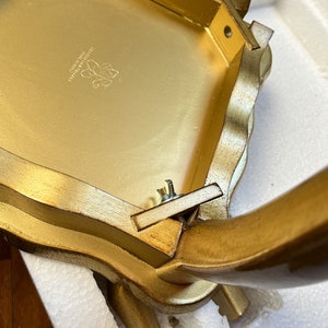 K&ouml;nnte beinhalten: Ein goldfarbener, verzierter Tisch mit gewelltem Rand. Die Unterseite des Tisches ist sichtbar und zeigt die Konstruktion und ein Logo. Die Beine sind gebogen und mit Schrauben befestigt. Die Website LaBallerinaGallery.com ist sichtbar.