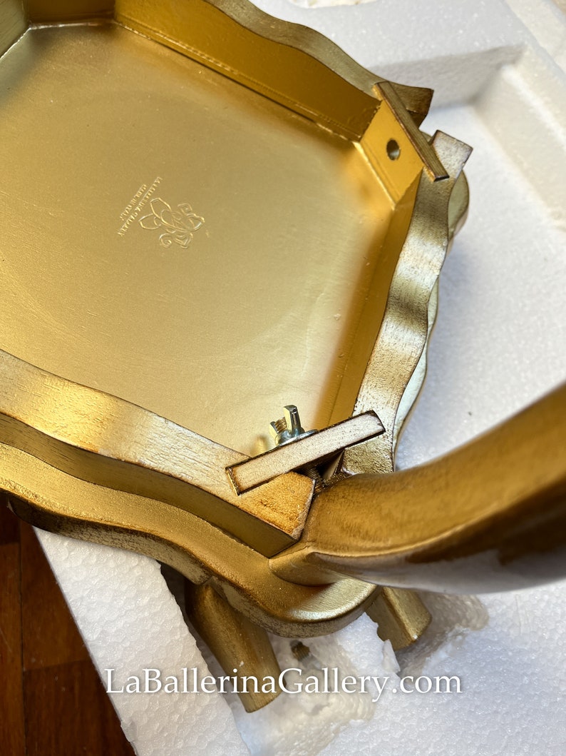 K&ouml;nnte beinhalten: Ein goldfarbener, verzierter Tisch mit gewelltem Rand. Die Unterseite des Tisches ist sichtbar und zeigt die Konstruktion und ein Logo. Die Beine sind gebogen und mit Schrauben befestigt. Die Website LaBallerinaGallery.com ist sichtbar.