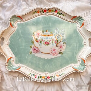 Puede incluir: Una bandeja de servir de cerámica blanca con un diseño decorativo de una tetera y flores sobre un fondo verde claro. La bandeja tiene un borde festoneado y un marco pintado en blanco y dorado.