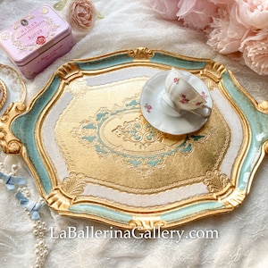 Puede incluir: Una bandeja decorativa dorada, blanca y turquesa con detalles ornamentados. Una taza de té blanca con detalles florales rosas se encuentra en la bandeja. La bandeja tiene un interior dorado y está rodeada por un borde turquesa. El sitio web LaBallerinaGallery.com es visible.