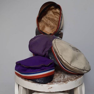 May include: Four vintage hats in a purple, white and red colour scheme. The hats have a flat brim and are made of a soft fabric. The hats are sitting on a white stool.