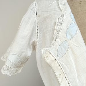 May include: A white blouse featuring short sleeves with lace trim and circular embroidery. The front placket includes buttons and a decorative collar edge with light blue oval accents and intricate lace detailing along the neckline and sleeve cuffs.