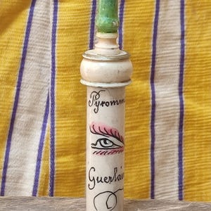 Peut inclure: Un flacon de parfum Guerlain vintage avec un bouchon vert. La bouteille cylindrique porte les mots "Pyromme" et "Guerlain". Une illustration d'un œil est également présente sur la bouteille.