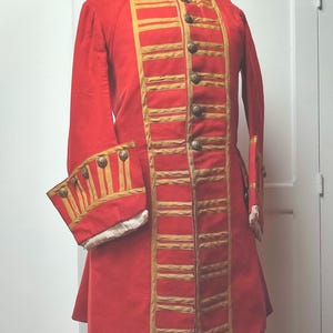 Puede incluir: Un abrigo rojo de estilo militar con adornos y botones dorados. El abrigo tiene un cuello alto y mangas largas con puños decorativos. El adorno dorado está dispuesto en bandas horizontales. El abrigo se exhibe en un maniquí.
