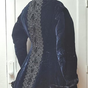 Puede incluir: Un abrigo vintage de terciopelo azul marino con adornos decorativos negros en la espalda y alrededor del cuello y los puños. El abrigo se exhibe en un maniquí, mostrando su diseño a medida y su lujosa textura.