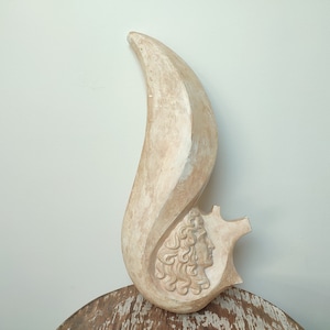 Może przedstawiać: Biała ceramiczna rzeźba przedstawiająca stylizowany płomień z wyrzeźbioną ludzką twarzą w profilu. Rzeźba jest abstrakcyjna i ma gładką, fakturowaną powierzchnię.