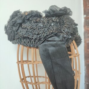 May include: A black hat with a large bow and sequins. The hat is on a wooden hat form.