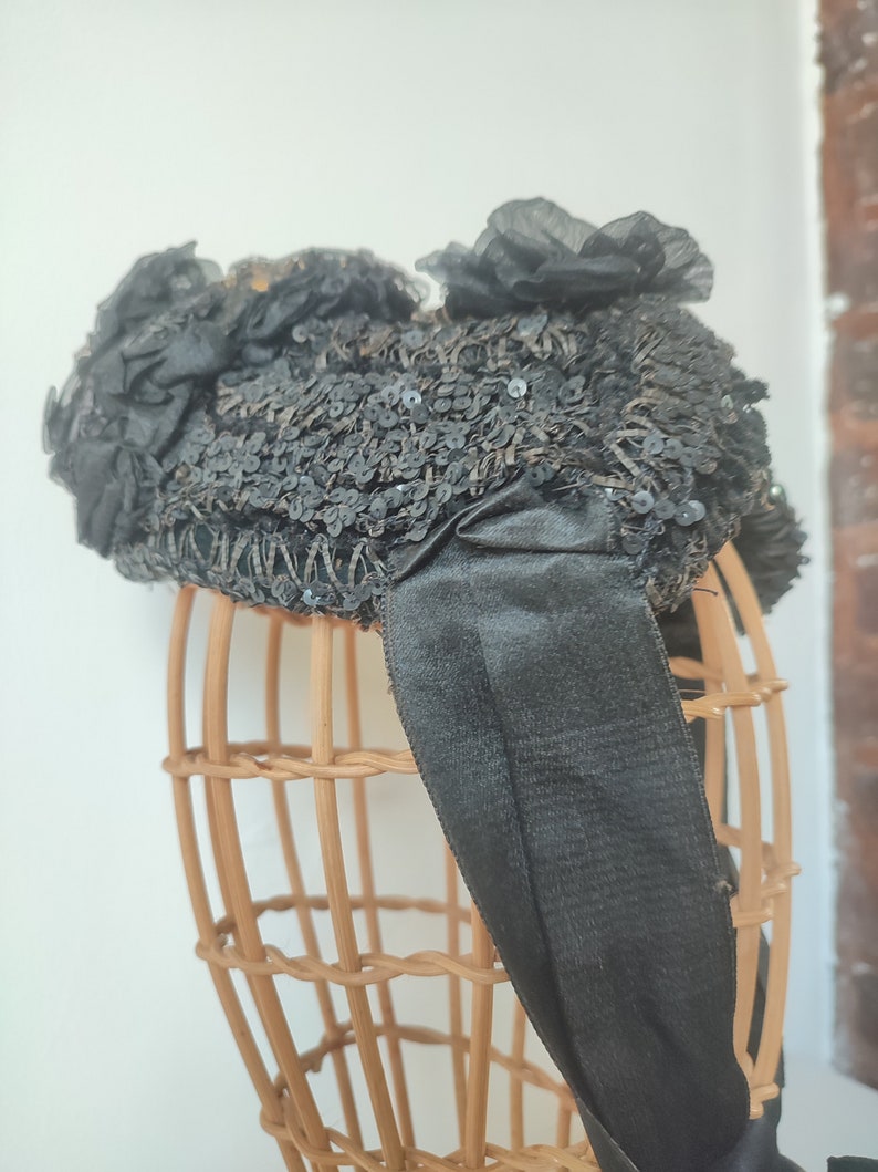 May include: A black hat with a large bow and sequins. The hat is on a wooden hat form.
