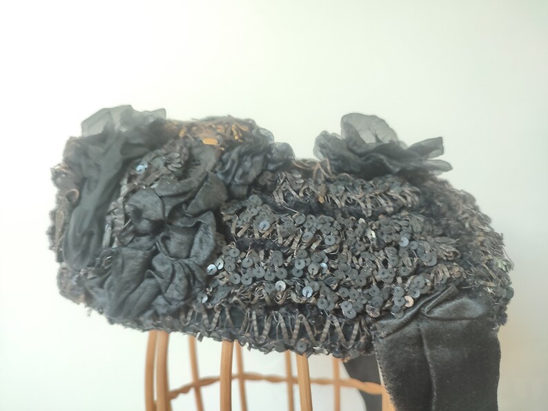 May include: A black hat with a large, ruffled bow and sequins. The hat is on a wooden hat stand.
