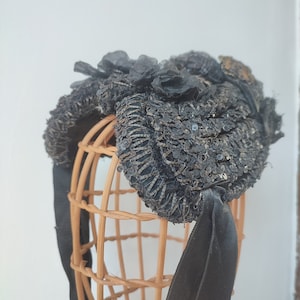 May include: A black sequined hat with a large black bow and floral embellishments. The hat is displayed on a wooden hat form.